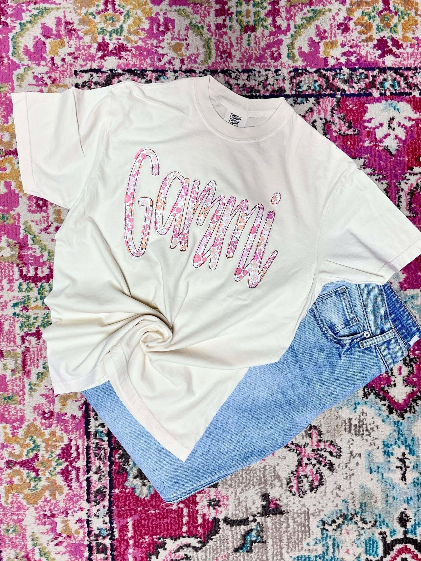 White t-shirt with colorful 'Gammi' text on a multicolored patterned surface