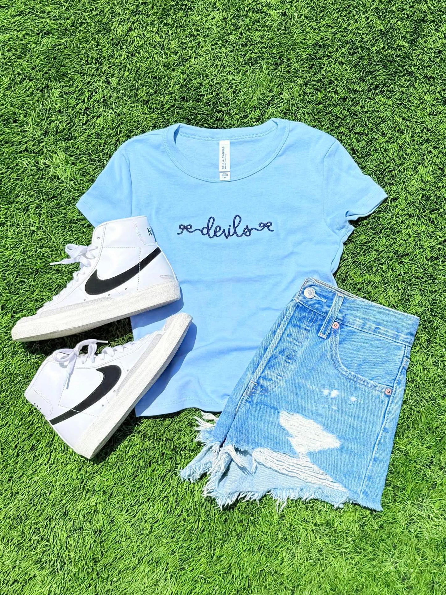 Light blue t-shirt with 'devils' text, white high-top sneakers, and denim shorts on grass