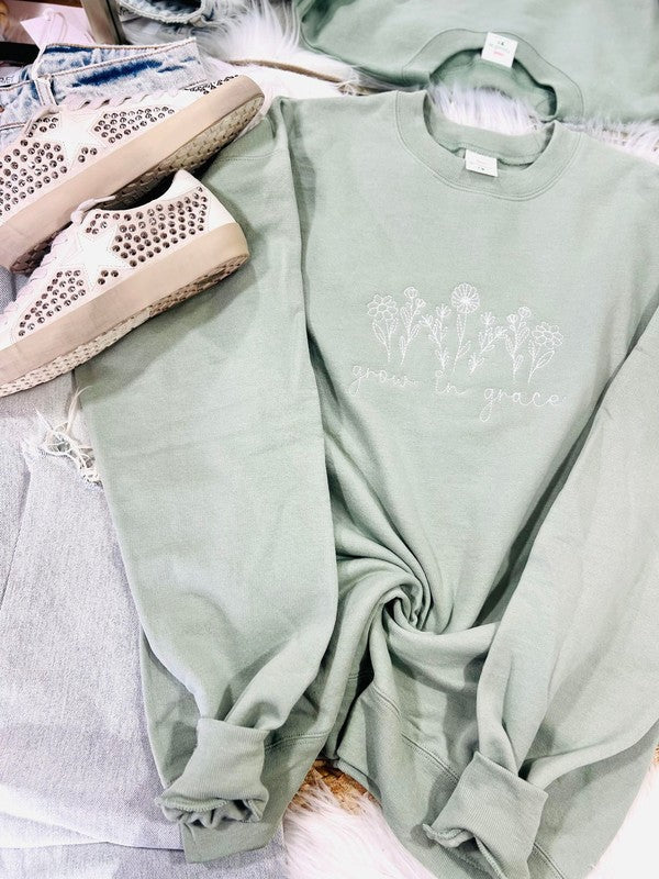 A green embroidered sweatshirt with 'wildflowers' text and a floral pattern, displayed flat with a pair of white sneakers and a denim jacket in the background.