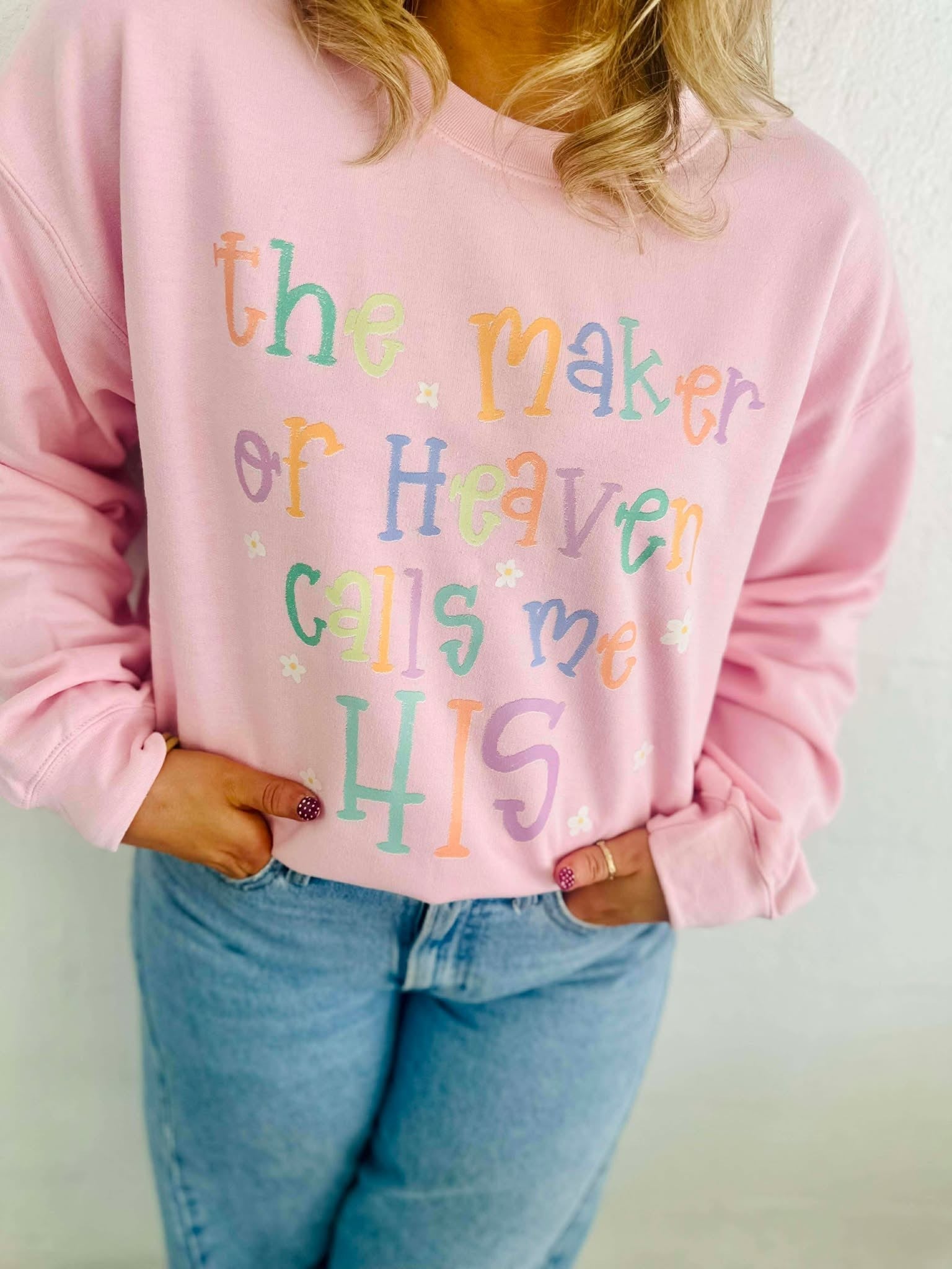 Person wearing a pink sweatshirt with colorful text design, the maker of heaven calls me HIS, standing against a plain background.