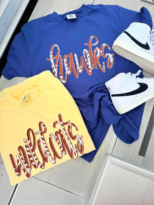 A flat-lay of two graphic t-shirts and a pair of white sneakers on a light gray tiled floor. The top blue t-shirt features 'hawks' in orange and white patterned script, and the bottom yellow t-shirt features 'wildcats' in a matching orange and white patterned script.